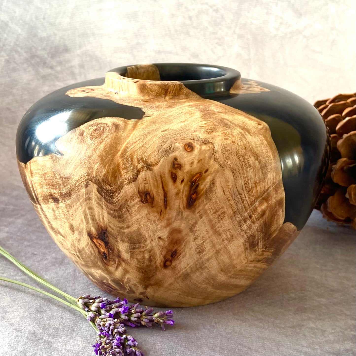 Wooden vase with black top on a gray surface with lavender and a pine cone.